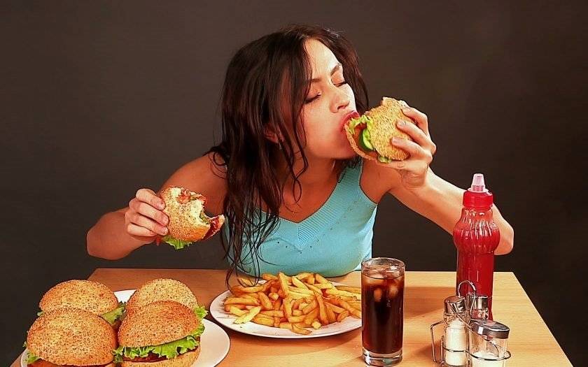 woman with long dark hair, binge eating, sitting at a wooden table, full of food, holding two burgers in her hands, taking a bite of one burger, burger and fries on the table, bottle and glass of coke in front of her // Healthier Me Today