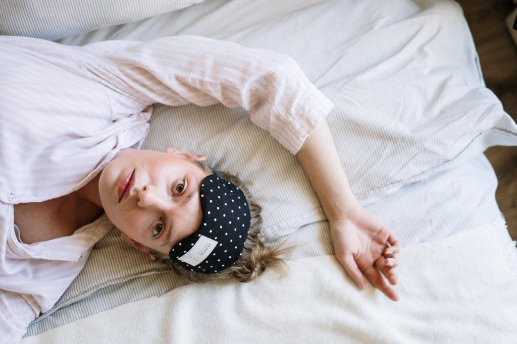 insomnia, woman lying on bed, white pajamas, black and white eye mask on head, hand above head // Healthier Me Today