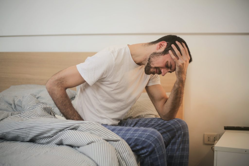 man wearing pajama's, sitting on bed, holding his head, wooden backboard // Healthier Me Today