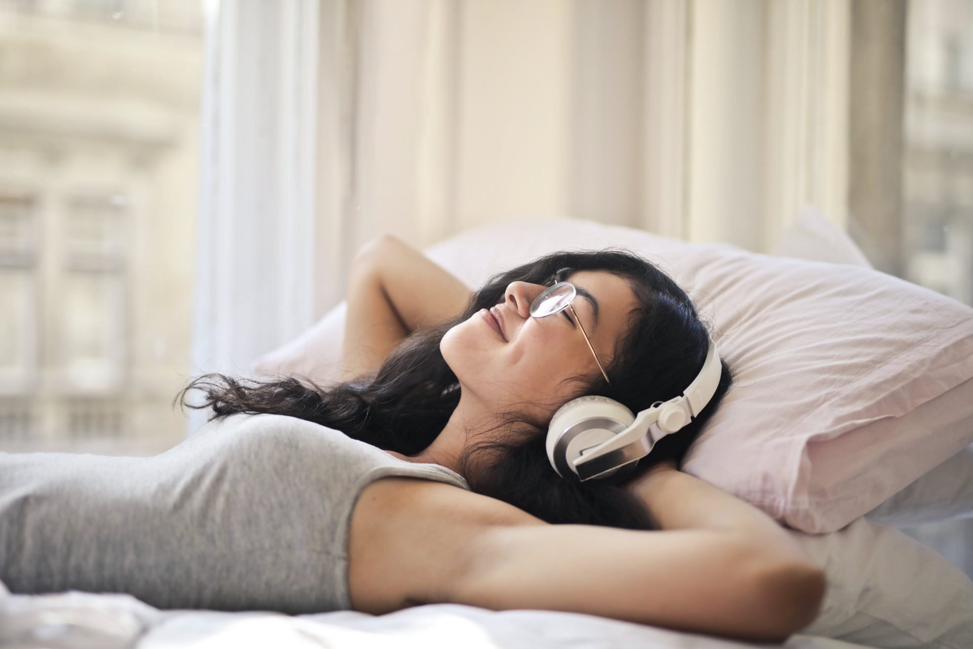 woman lying on bed, smiling, listening to music through headphones, white headphones, wearing grey tank top, wearing glasses // Healthier Me Today