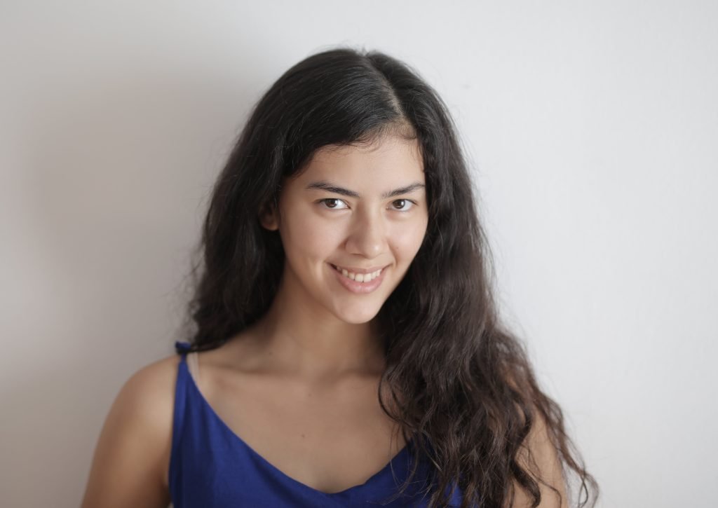 woman with long curly black hair, smiling, wearing a blue top, standing against white wall // Healthier Me Today