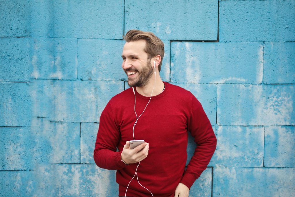 man standing, smiling, wearing red long shirt, white cabled headphones in ears, blue wall // Healthier Me Today