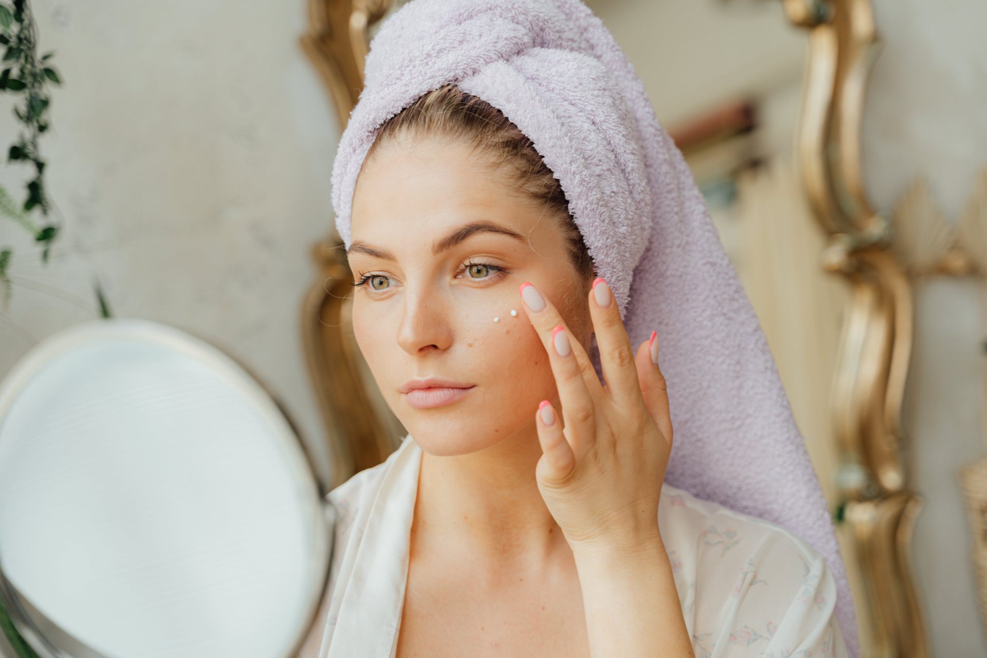 woman in bath robe, hair wrapped in towel, dark hair, doing skincare, looking at mirror, blurred background, gold mirror behind woman // Healthier Me Today