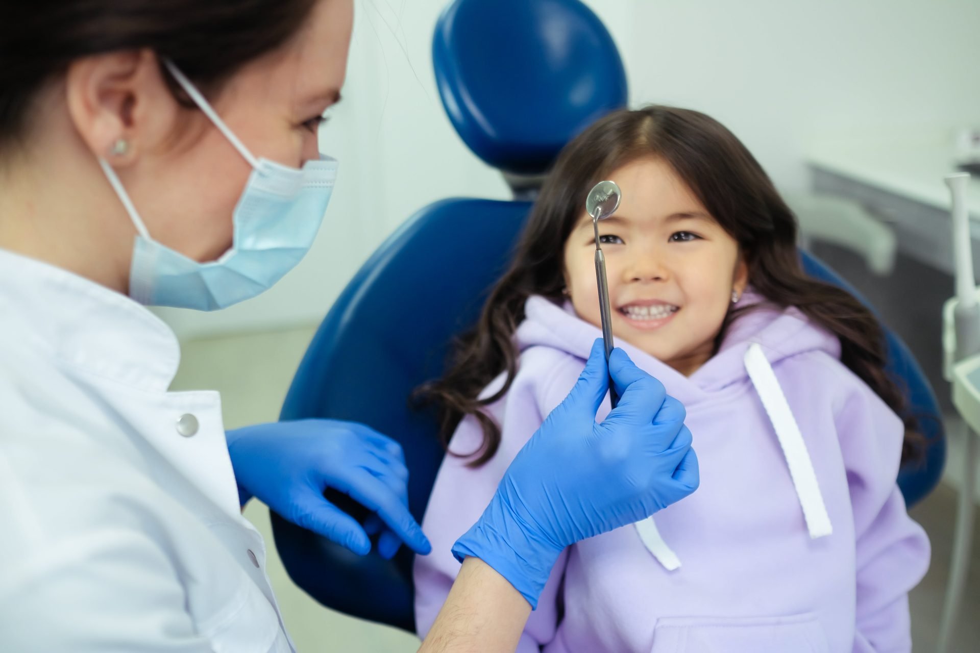 child with dark long hair, smiling, sitting on dentist chair, dentist with mask looking at child's teeth // Healthier Me Today