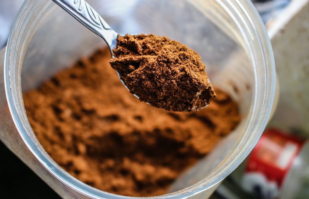 muscle recovery supplements: close shot of container, brown protein power, heaped teaspoon. blurred background // Healthier Me Today