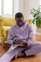 man sitting on floor, looking at magazine, wearing purple set of sweats, yellow chair, plant on table, big window // Healthier Me Today